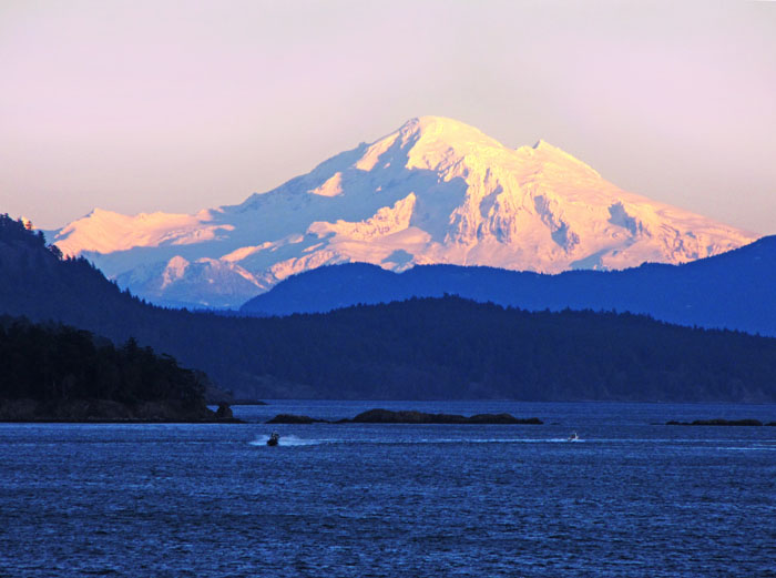 Photo of Mt Baker, Washington State at sunset, taken from Vancouver Island, with a speedboat on the foreground water coming toward the camera.