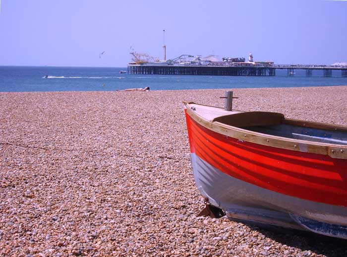 Photo of a sunny beach with a red boat in the foreground a woman sunbathing in the mid and a pier and boat on the sea in the bachground
