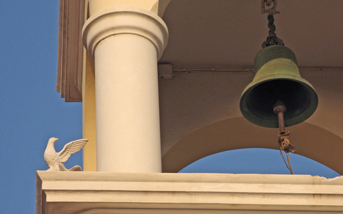 A white dove next to a bell tower looking and pointing with it's wing at the bell